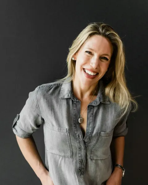 Portrait of Dawn Barry, laughing, in a grey button-up shirt against a dark wall